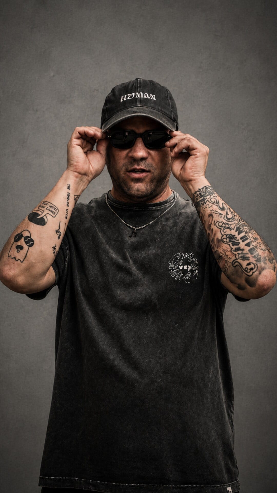 Tomas Tassara in a black cap and washed black tee against a grey backdrop, lowering his sunglasses towards the camera, tattooed forearms raised.